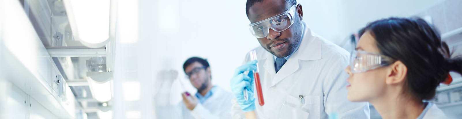 Female Asian laboratory scientist in lab coat and safety goggles showing test tube with red liquid to curious African-American colleague in laboratory  Latin-American scientist in background 
