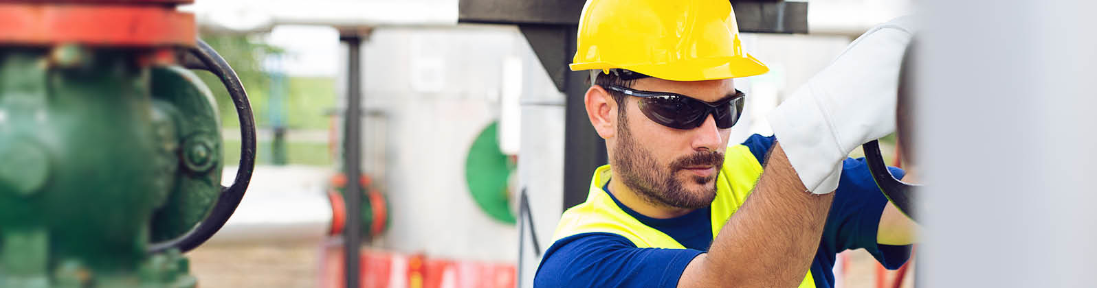 Man wearing sunglasses, safety vest, and hard hat working on outdoor plant pipes 